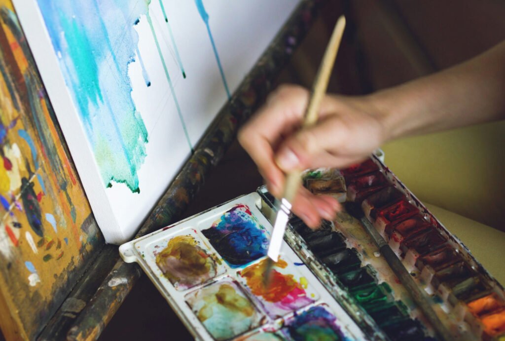Hand painting with watercolour beside a canvas, suggesting process-led work for a Parsons BFA Fine Arts portfolio.