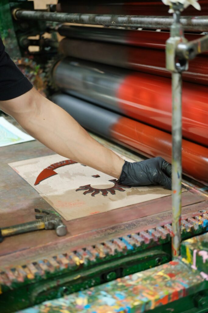 Gloved hand working at a printing press with red-inked rollers, for applicants meeting the Yale Painting Printmaking MFA portfolio requirements