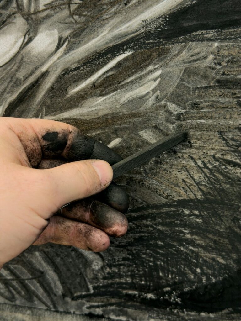 Charcoal-blackened hand holding a stick of charcoal over a dark, heavily worked drawing, for applicants preparing a SAIC Painting and Drawing portfolio