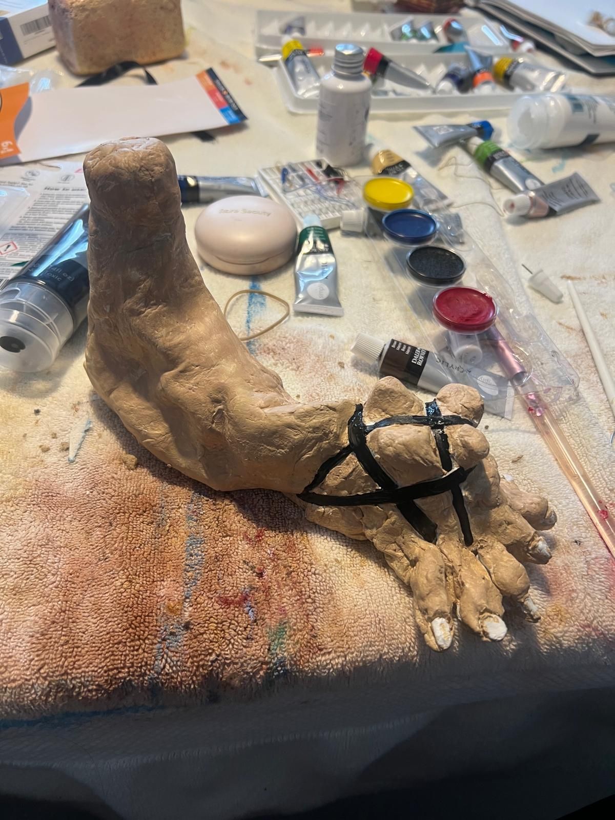 Work in progress: a sculpted foot with black straps on a table covered in paint tubes and brushes